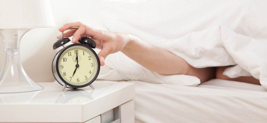 Hand under blanket reaching out for alarm clock, shallow depth of field focus on foreground